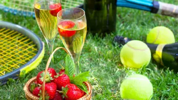 Close up of two glasses of champagne, a basket of strawberries, tennis balls and rackets on some grass