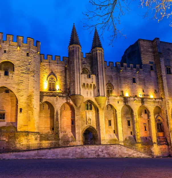 Low angle shot of a beige coloured stone palace, lit up with amber lights on the front. With pointy turrets in the middle and battlements on the left side. In front of a bright blue night sky.