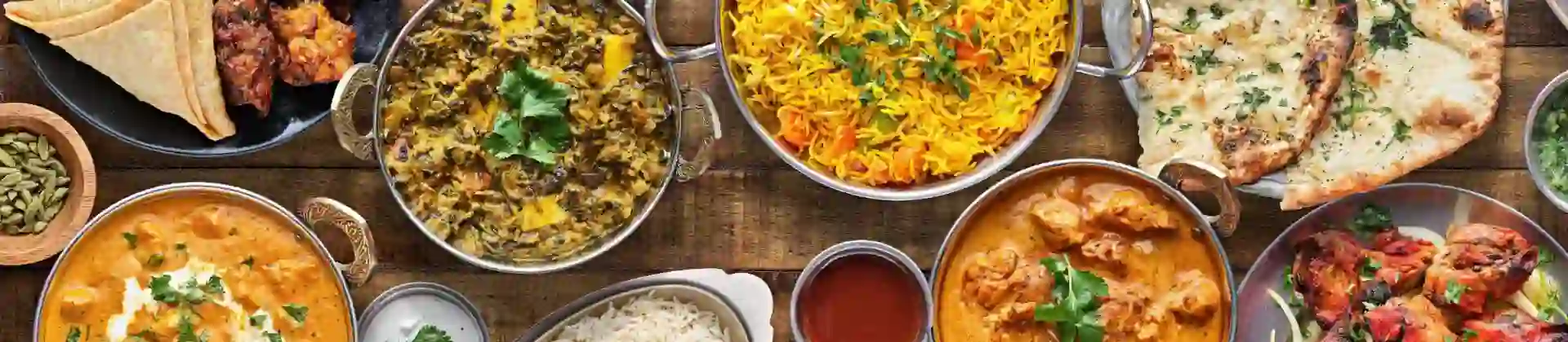 A colourful spread of Indian food featuring dishes such as curry, naan bread, rice, samosas, and chutneys served in bowls and plates on a wooden table