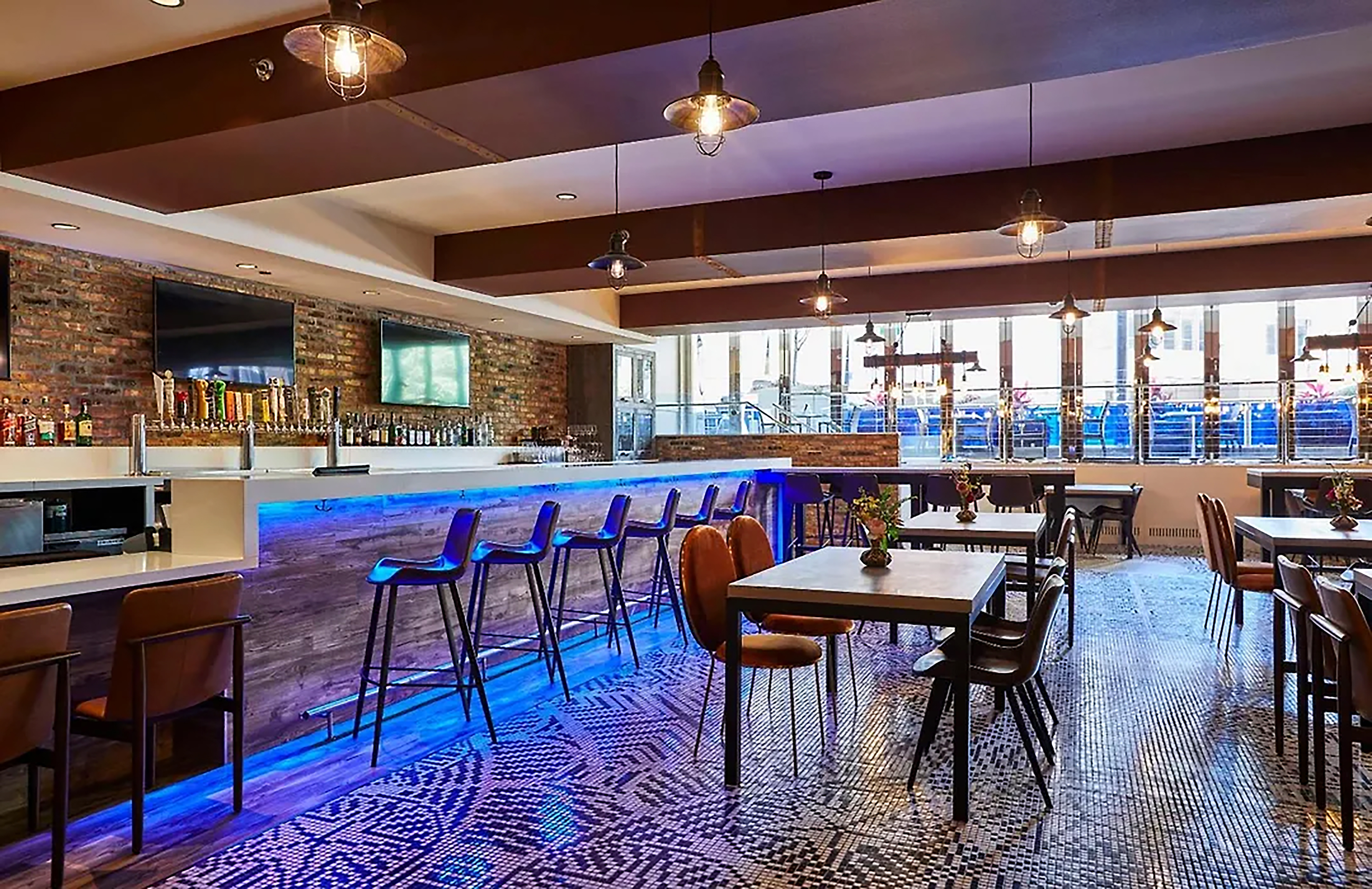 Bar at Radisson Blu Aqua Hotel, Chicago, featuring blue under-bar lighting, dim ambiance with warm ceiling lights and dark, modern furnishings and tables and chairs arranged around the space