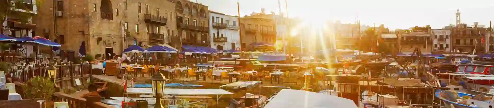 View of a marina from a restaurant, with a laid table in the forefront behind a wooden barrier dividing the boats on the water and the land. Other buildings and restaurants can be seen on the other side, with blue umbrellas and outdoor seating. The sun is poking over the buildings, with beams shining into the camera