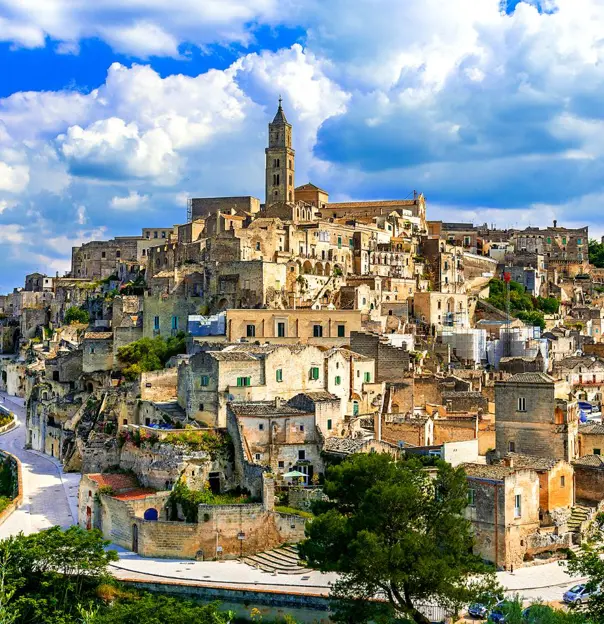 A panoramic view of the historic stone town of Matera, Italy, with ancient buildings stacked on a hillside under a partly cloudy blue sky
