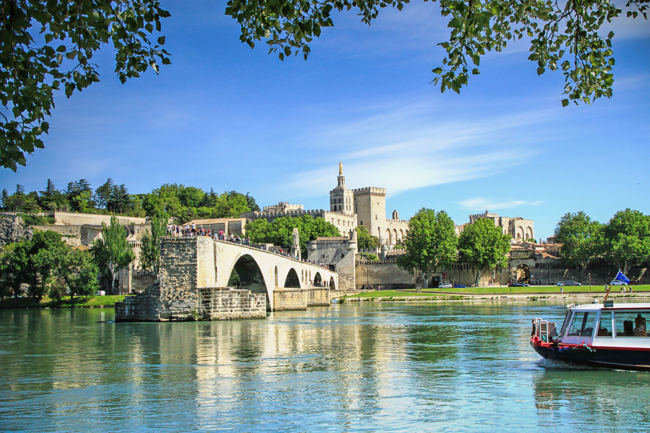View of the Pont D’Avignon bridge, with the river water in the forefront and a boat coming from the right. In the distance, on the other side of the bridge is a grey palace. The sky is blue.