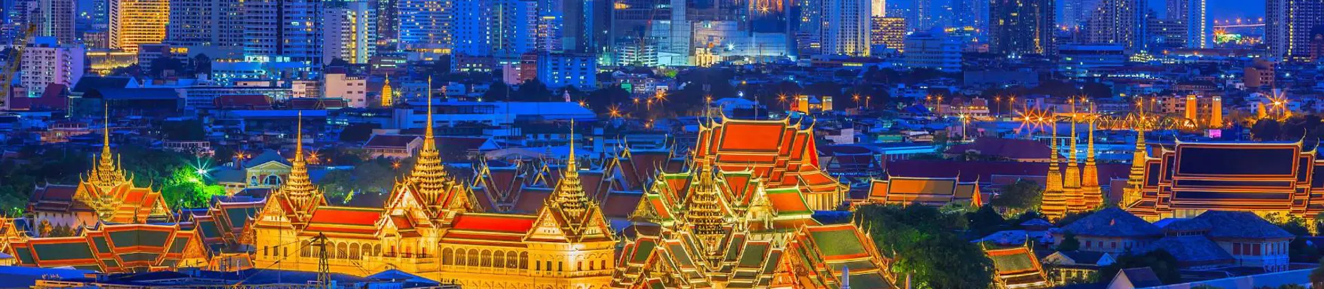 Distant view of the Grand Palace in Bangkok lit up at night, with its golden spires and ornate rooftops surrounded by softly illuminated temples and skyscrapers