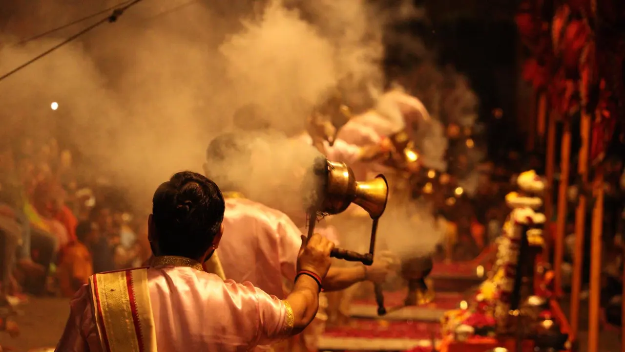 A close view of priests performing the Ganga Aarti ritual in Varanasi, holding brass lamps with incense smoke swirling in the air amid a reverent evening crowd