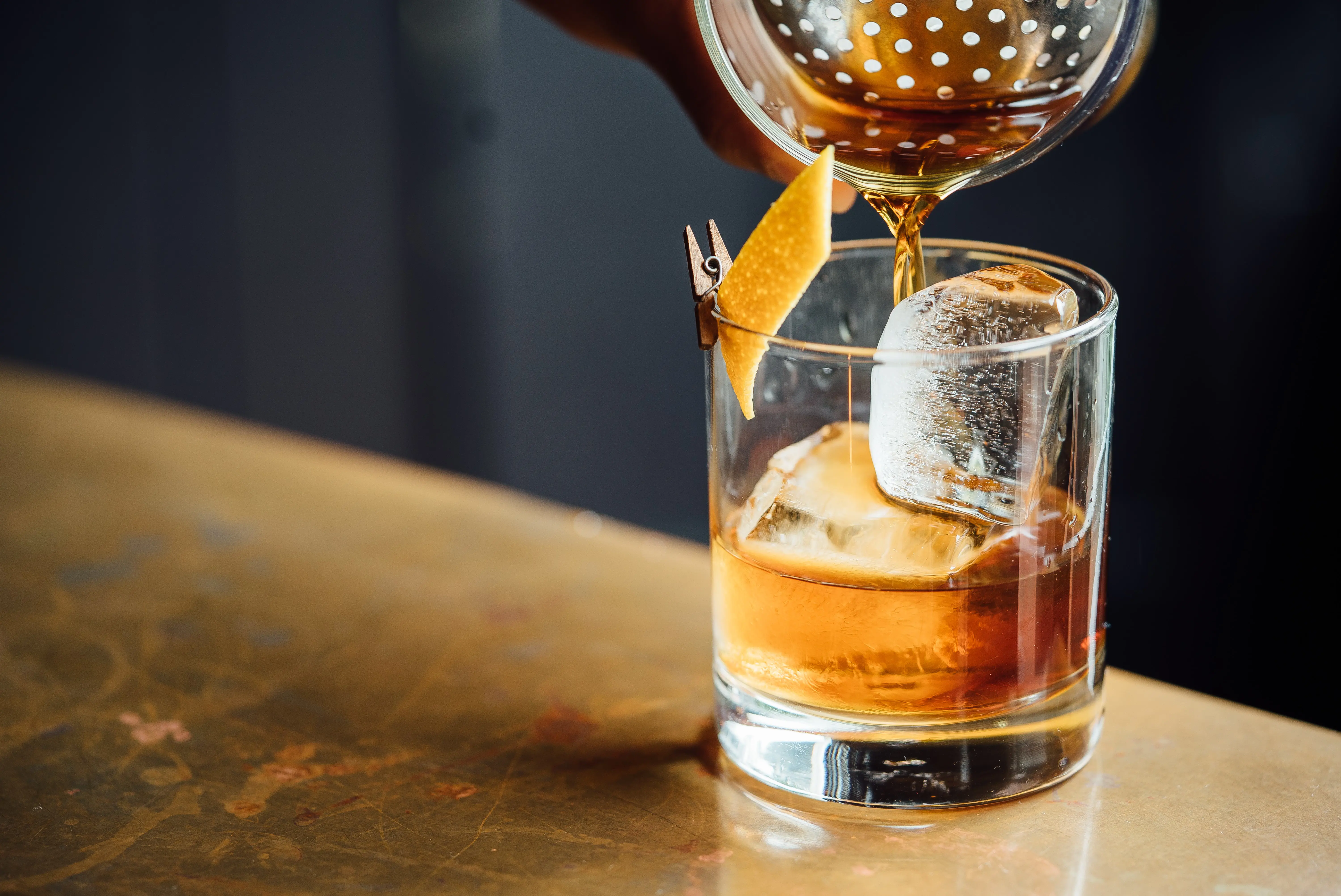 Close up on a whisky being poured, with ice in the glass and lemon peel clipped to the side of the glass. 