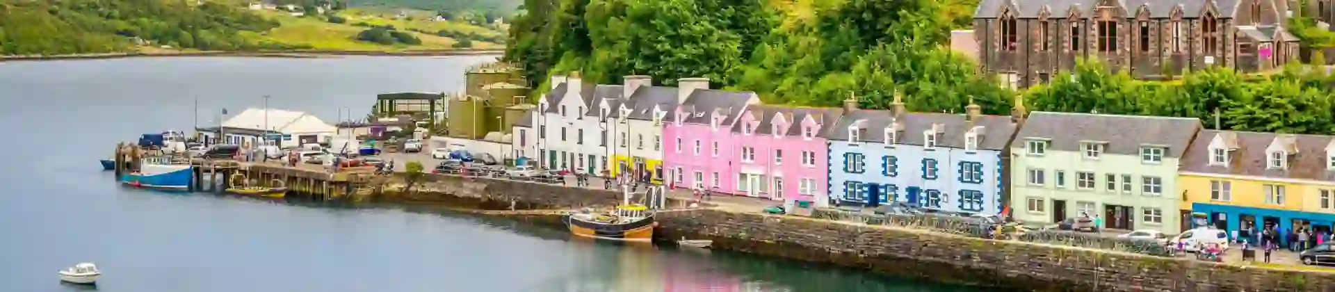 The colourful harbourfront of Portree on the Isle of Skye in Scotland, with pastel-painted houses reflected in the calm water, nestled beneath a lush green hill on a sunny day