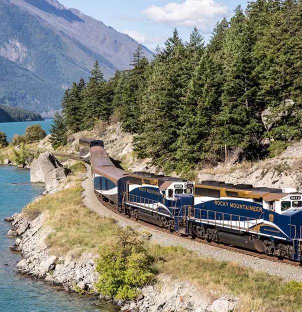 The Rocky Mountaineer train travelling through Canada beside a tranquil lake, with tall trees and scenic mountain views in the background