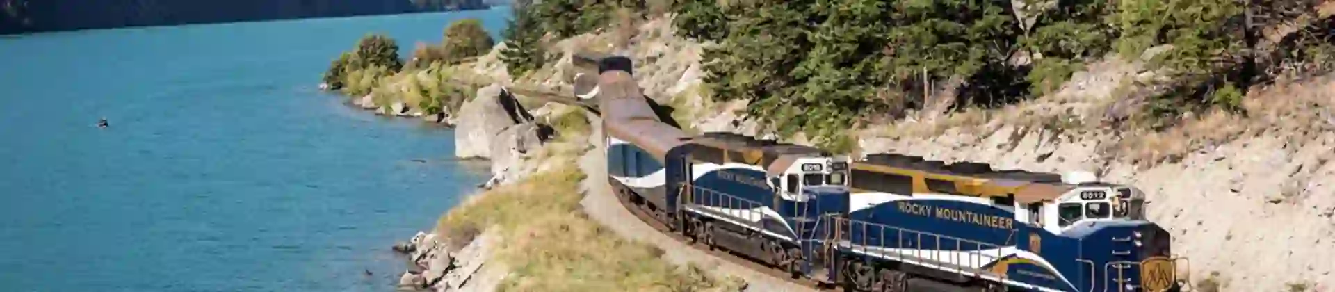 The Rocky Mountaineer train travelling through Canada beside a tranquil lake, with tall trees and scenic mountain views in the background