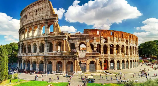 The Colosseum in Rome, a vast ancient Roman amphitheatre with partially preserved stone arches and tiered seating, set against a blue sky with scattered clouds