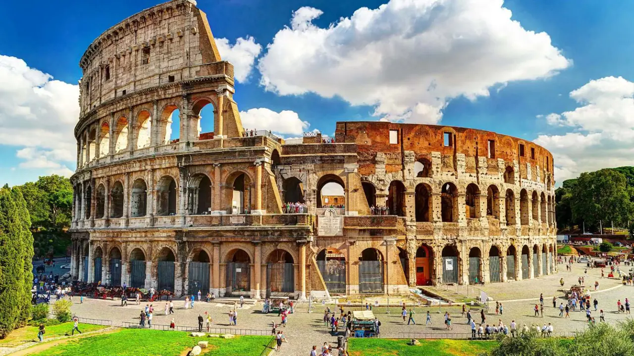 The Colosseum in Rome, a vast ancient Roman amphitheatre with partially preserved stone arches and tiered seating, set against a blue sky with scattered clouds