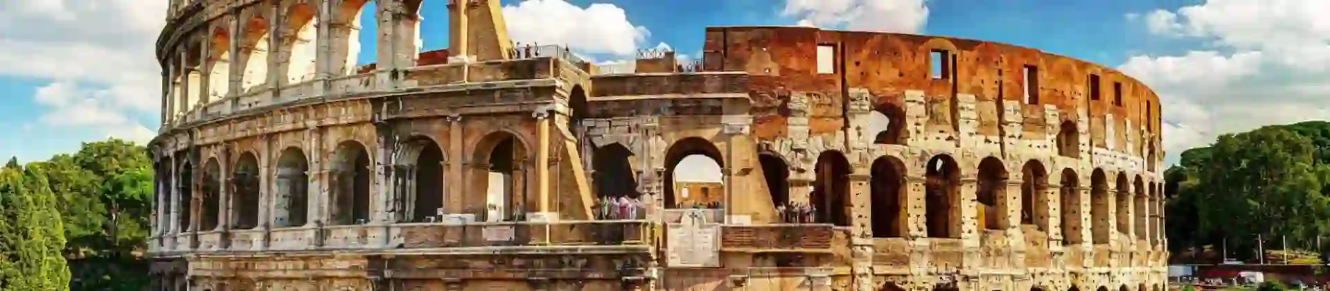 The Colosseum in Rome, a vast ancient Roman amphitheatre with partially preserved stone arches and tiered seating, set against a blue sky with scattered clouds