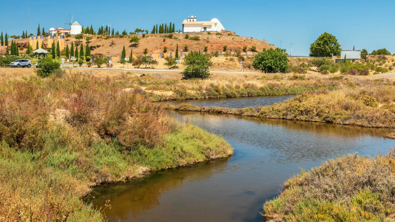 Picturesque Castro Marim Nature Reserve in Portugal, featuring a small river flowing beside shrubland, with a whitewashed windmill and house on the hilltops in the distance