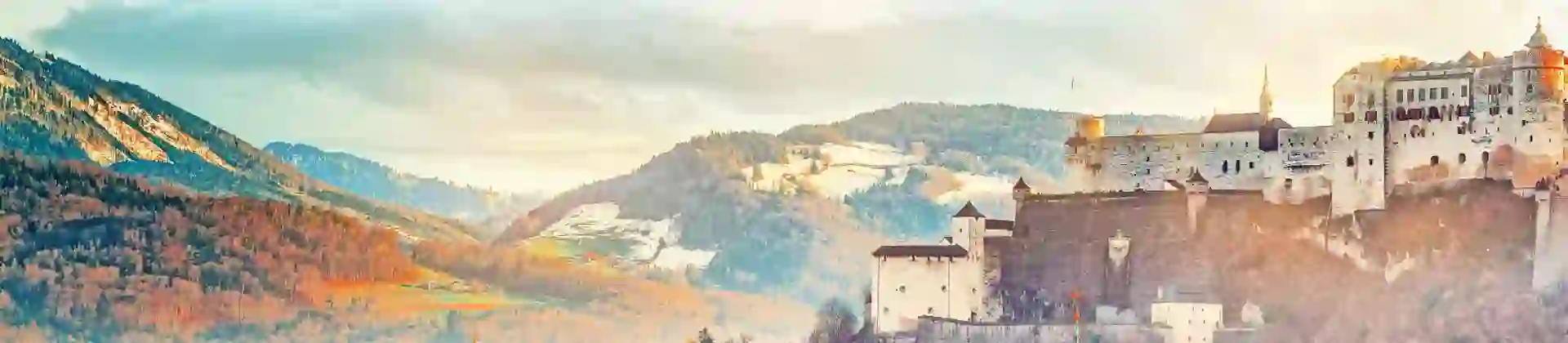 Shot of Salzburg, showing the top half of the city's buildings, with turquoise turrets dotted around the image and a wide church in the forefront and a cathedral behind it. Mountainous landscape behind and the fortress on the hill to the right.