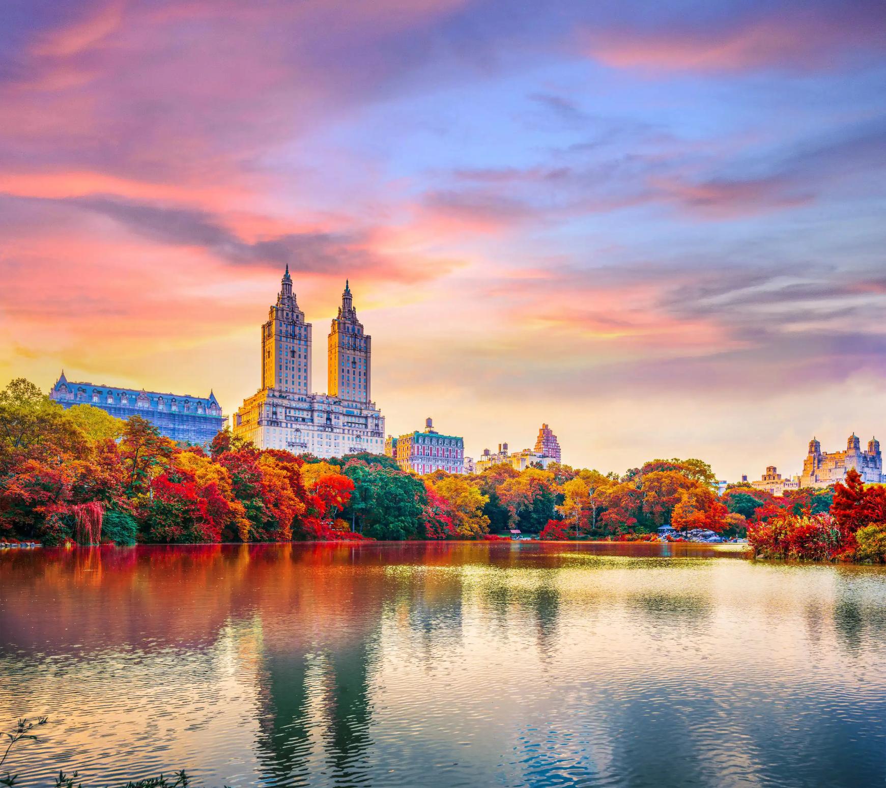 Central Park in autumn with The Lake in the centre, surrounded by colourful trees in shades of red, orange, green and yellow. The iconic twin towers of The San Remo rise in the background