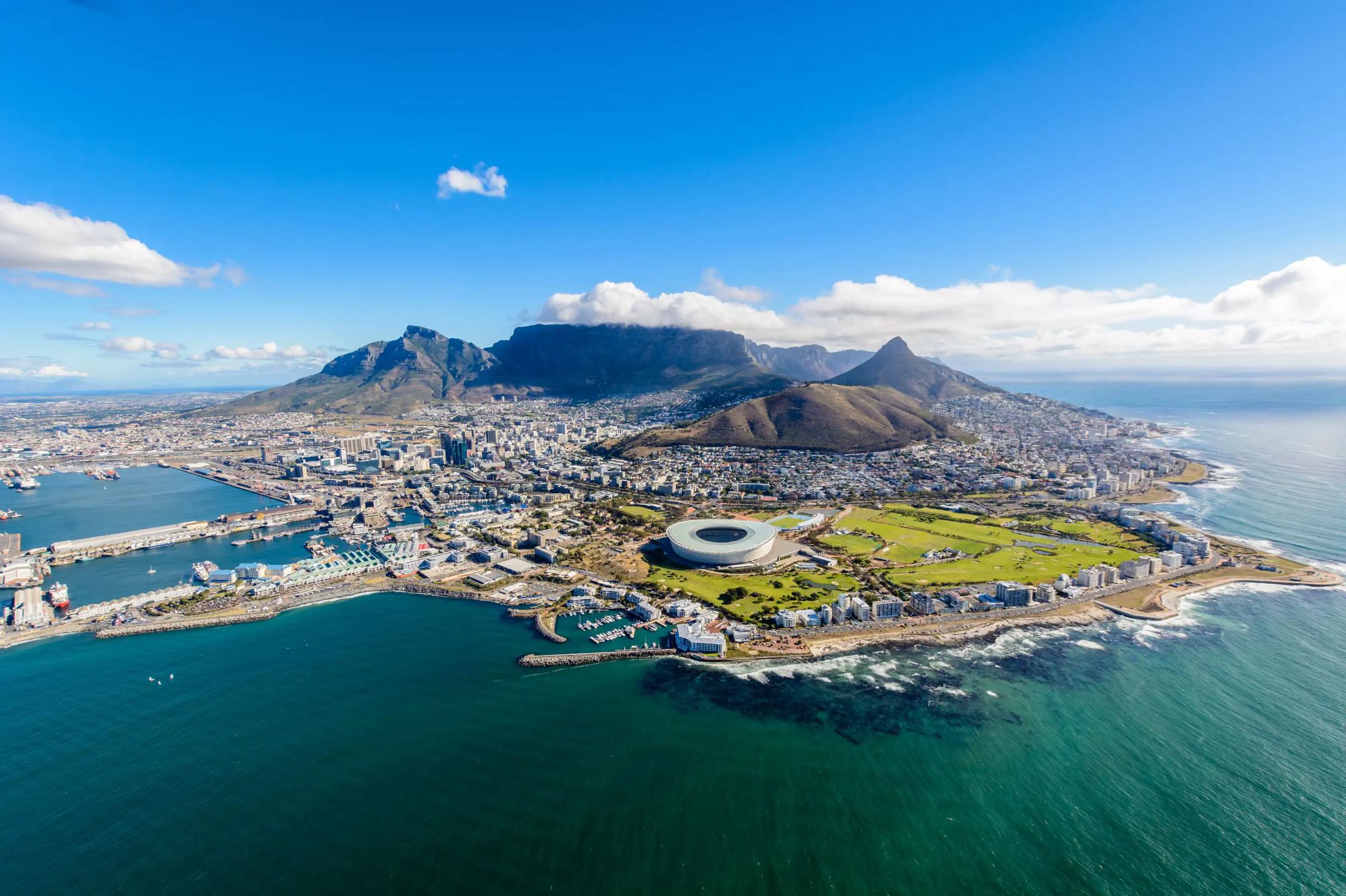 Aerial view of Cape Town, South Africa, showing the city nestled between Table Mountain and the Atlantic Ocean, with the coastline, harbour, and urban landscape clearly visible