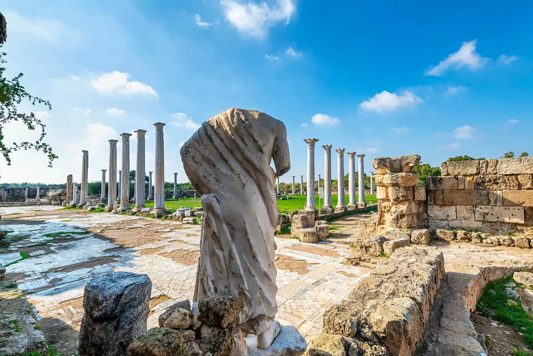 View of Roman ruins in Salamis, with the back of a headless marble statue. Further away, there is a green lawn surrounded by marble pillars