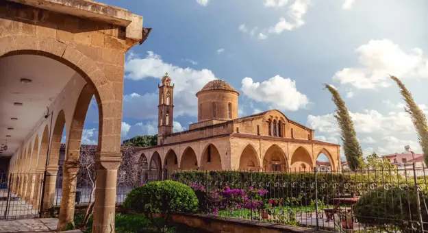 A stone Greek Orthodox monastery with arched walkways, a domed roof, and a bell tower. A well-kept garden with flowers and shrubs lines the foreground under a sunny sky