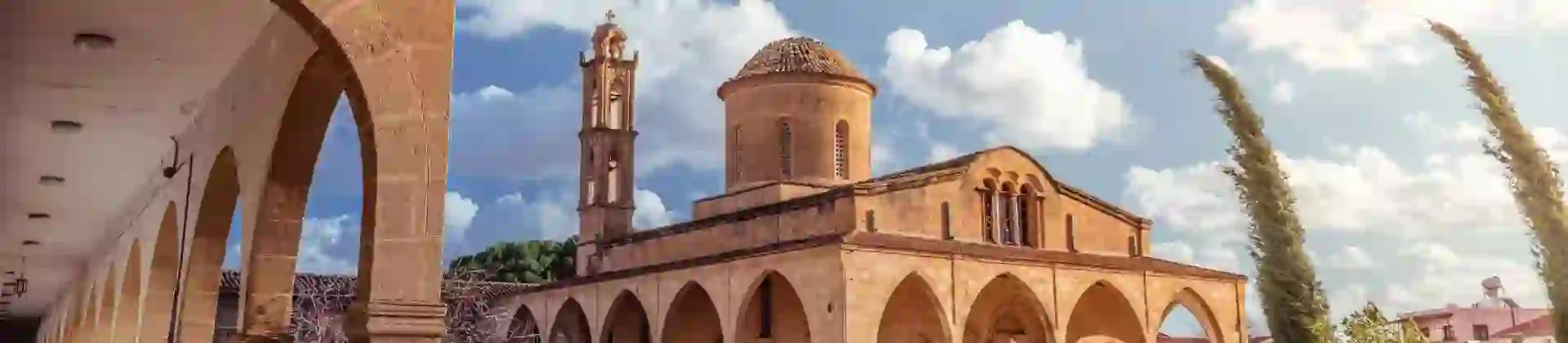 A stone Greek Orthodox monastery with arched walkways, a domed roof, and a bell tower. A well-kept garden with flowers and shrubs lines the foreground under a sunny sky