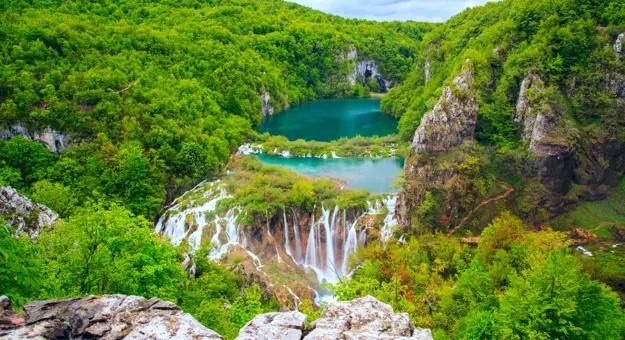 High shot of waterfalls in front of a dark turquoise body of water, surrounded by forested hills. In the forefront is a piece of rock and trees.