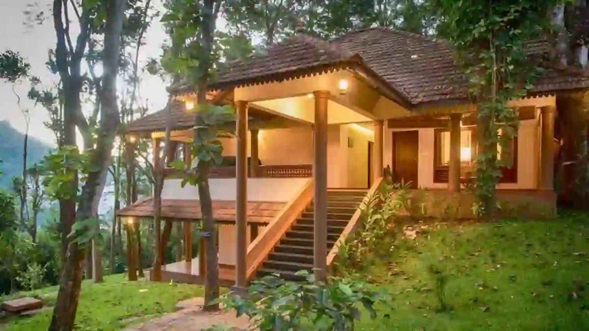 Exterior view of Greenwoods Hotel in Periyar, nestled among trees and greenery, with warm lighting highlighting the traditional building as dusk falls