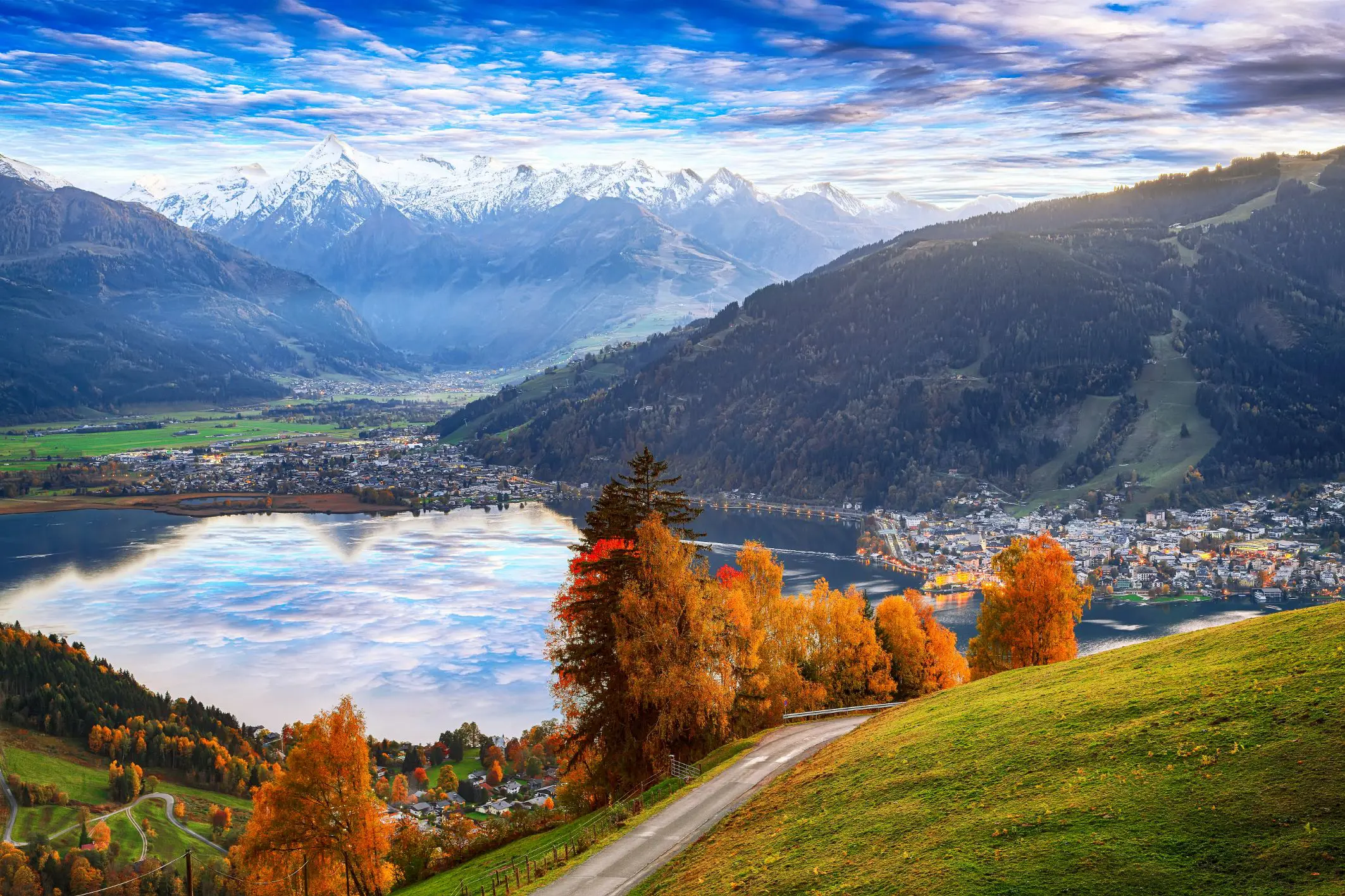 View of Austrian mountains, with some in the distance with snow on the top. Below is a town where small buildings can be seen, behind a lake. To the right another town, and in the forefront, orange autumnal trees and a grassy hill and a pathway
