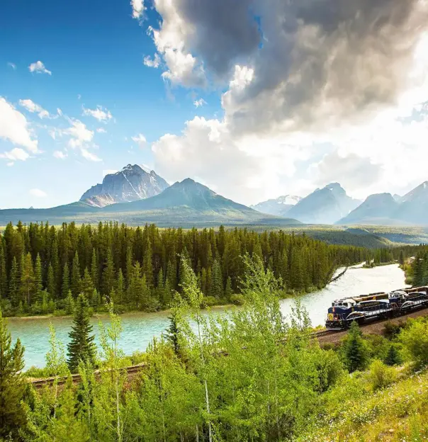 A train travelling through the Canadian Rockies, with rugged mountains in the background and dense evergreen forests lining the tracks