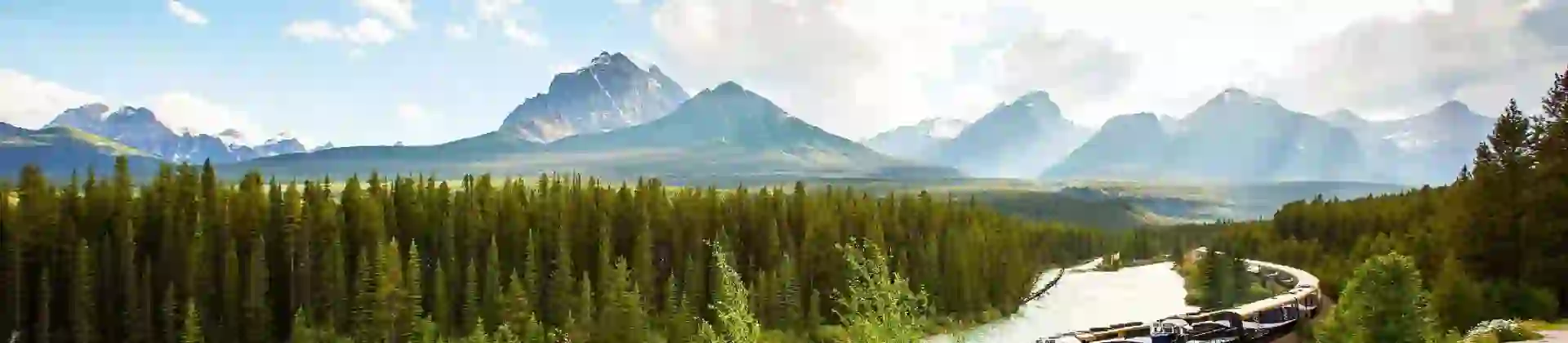 The Rocky Mountaineer train travelling through the Canadian Rockies, with rugged mountains in the background and dense evergreen forests lining the tracks